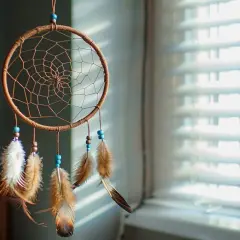 Attrape-rêves artisanal avec plumes et perles bleues, suspendu devant des stores.