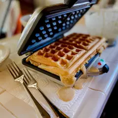 Gaufrier ouvert avec une gaufre dorée. De la pâte déborde. Des fourchettes attendent. Pensez à comment nettoyer un gaufrier après ce délice !