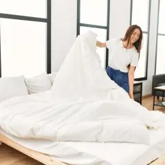 Une femme souriante apprend comment mettre une housse de couette à l'envers, en la drapant sur un lit blanc.