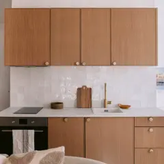 Petite cuisine de studio avec placards en bois, carrelage blanc et lit en arrière-plan.
