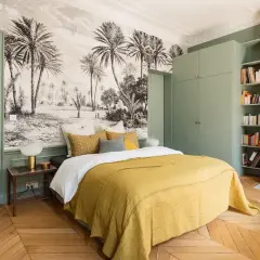 Une chambre adulte avec un papier peint paysage tropical, un lit jaune, une armoire verte et une cheminée.
