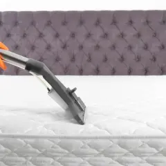Main tenant en orange nettoie un matelas blanc avec un appareil à vapeur. Le nettoyage du matelas à la vapeur est essentiel pour une literie saine.