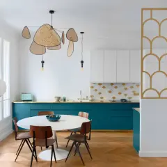 Cuisine moderne avec des armoires bleu canard, un dosseret à motifs dorés et blancs, et une table ronde. La composition de cuivre blanc se retrouve dans le luminaire aérien et la séparation décorative.