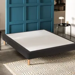 Un sommier tapissier noir aux pieds en bois, prêt à accueillir un matelas. Il est posé sur un tapis géométrique.