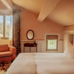 Chambre avec lit blanc, fauteuil orange, miroir rond et fenêtres donnant sur la verdure.