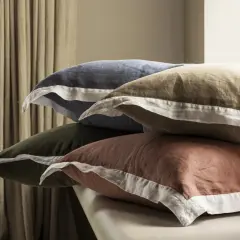 Pile de taies d'oreiller de différentes couleurs (bleu, kaki, marron) avec passepoil blanc.