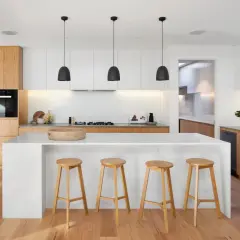 Cuisine moderne avec îlot central, tabourets en bois et éléments de **création cuisine** épurés. Éclairage suspendu noir et escalier adjacent.