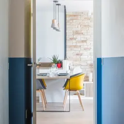 Vue d'un couloir avec une peinture bicolore bleu et blanc. Une porte s'ouvre sur une salle à manger moderne avec une table dressée et des chaises colorées.