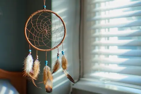 Attrape-rêves artisanal avec plumes et perles bleues, suspendu devant des stores.