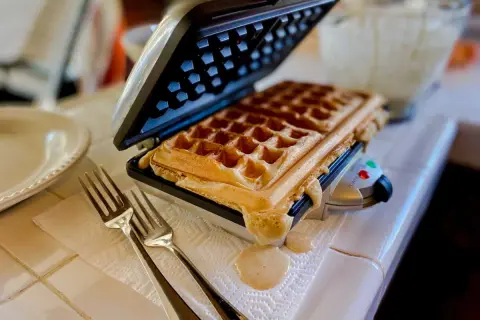Gaufrier ouvert avec une gaufre dorée. De la pâte déborde. Des fourchettes attendent. Pensez à comment nettoyer un gaufrier après ce délice !
