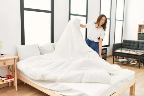 Une femme souriante apprend comment mettre une housse de couette à l'envers, en la drapant sur un lit blanc.