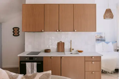 Petite cuisine de studio avec placards en bois, carrelage blanc et lit en arrière-plan.