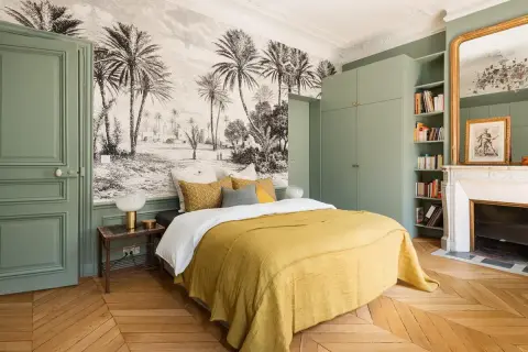 Une chambre adulte avec un papier peint paysage tropical, un lit jaune, une armoire verte et une cheminée.