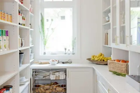 Arrière-cuisine blanche avec étagères remplies, fruits et légumes sur le comptoir.