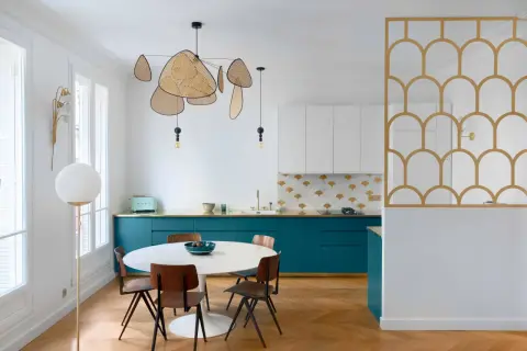 Cuisine moderne avec des armoires bleu canard, un dosseret à motifs dorés et blancs, et une table ronde. La composition de cuivre blanc se retrouve dans le luminaire aérien et la séparation décorative.