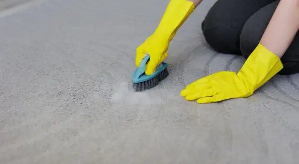 Mains gantées de jaune frottent un tapis gris avec une brosse, montrant comment nettoyer tapis.