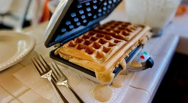 Gaufrier ouvert avec une gaufre dorée. De la pâte déborde. Des fourchettes attendent. Pensez à comment nettoyer un gaufrier après ce délice !