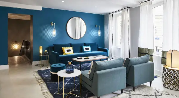 Salon moderne aux murs bleu paon, avec canapé velours, fauteuils assortis, tapis graphique et éclairage tamisé.