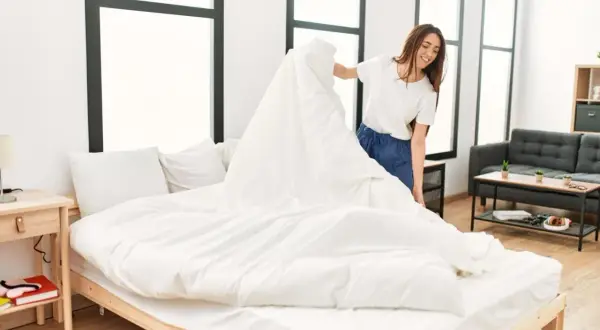 Une femme souriante apprend comment mettre une housse de couette à l'envers, en la drapant sur un lit blanc.