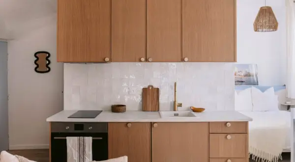 Petite cuisine de studio avec placards en bois, carrelage blanc et lit en arrière-plan.
