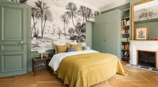 Une chambre adulte avec un papier peint paysage tropical, un lit jaune, une armoire verte et une cheminée.