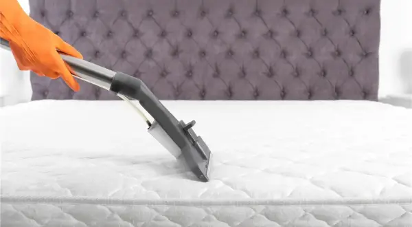 Main tenant en orange nettoie un matelas blanc avec un appareil à vapeur. Le nettoyage du matelas à la vapeur est essentiel pour une literie saine.
