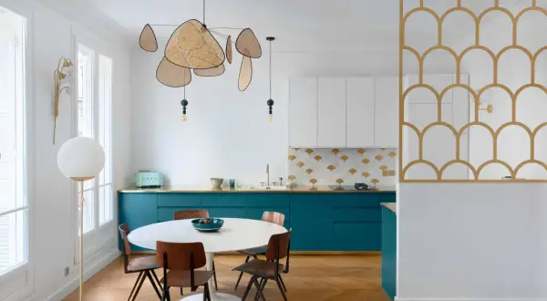 Cuisine moderne avec des armoires bleu canard, un dosseret à motifs dorés et blancs, et une table ronde. La composition de cuivre blanc se retrouve dans le luminaire aérien et la séparation décorative.