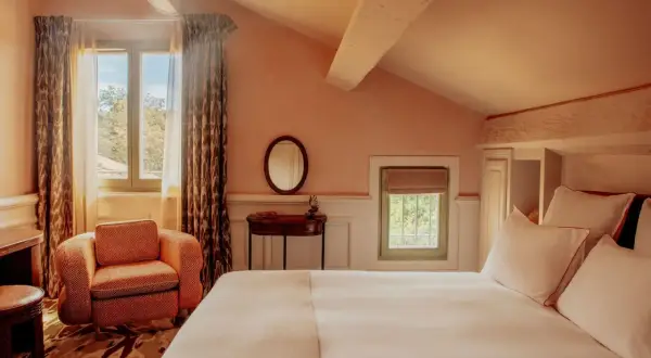 Chambre avec lit blanc, fauteuil orange, miroir rond et fenêtres donnant sur la verdure.