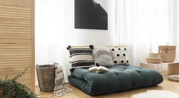 Futon vert foncé avec coussins décoratifs, panier en osier et tableau abstrait.