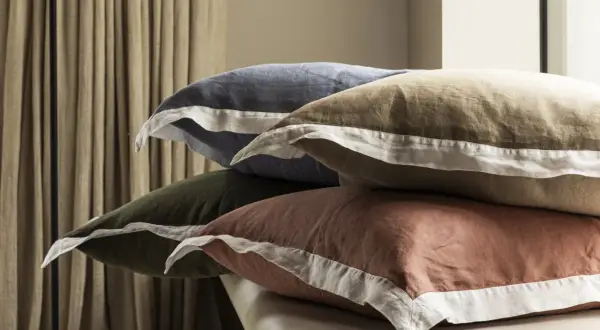 Pile de taies d'oreiller de différentes couleurs (bleu, kaki, marron) avec passepoil blanc.