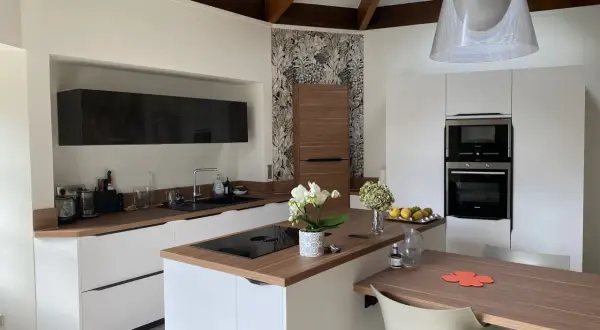Cuisine moderne avec des meubles blancs et un **crédence et plan de travail assortis** en bois. Une orchidée blanche décore l'îlot central.