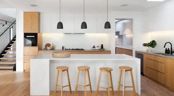 Cuisine moderne avec îlot central, tabourets en bois et éléments de **création cuisine** épurés. Éclairage suspendu noir et escalier adjacent.