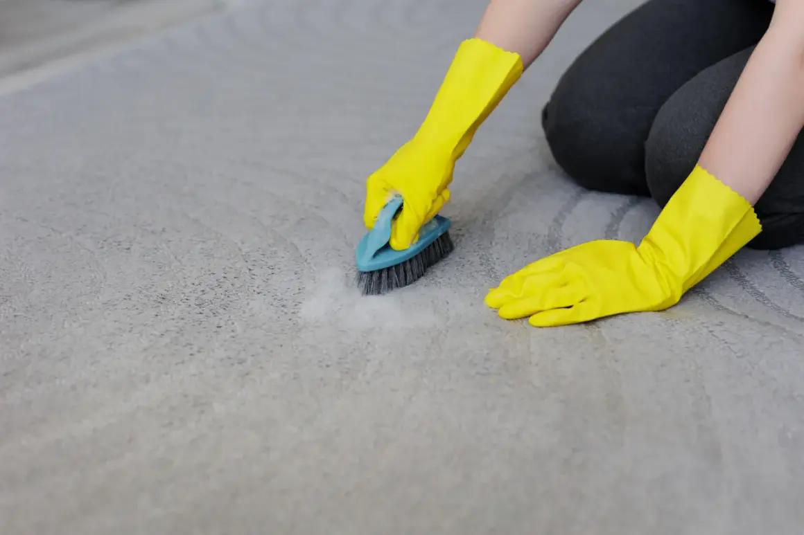 Mains gantées de jaune frottent un tapis gris avec une brosse, montrant comment nettoyer tapis.