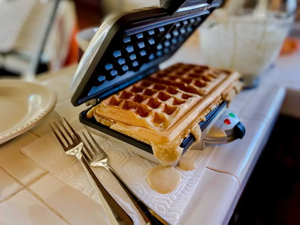 Gaufrier ouvert avec une gaufre dorée. De la pâte déborde. Des fourchettes attendent. Pensez à comment nettoyer un gaufrier après ce délice !