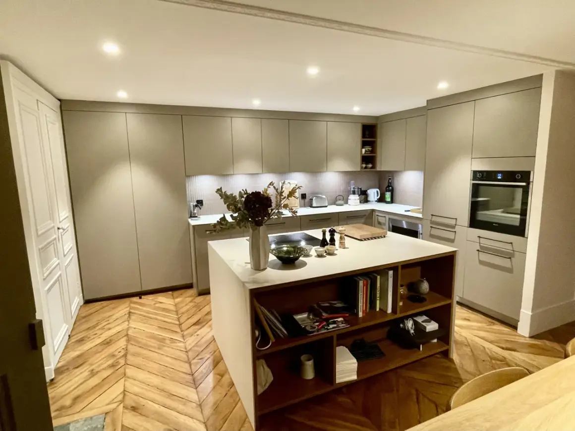 Cuisine moderne avec îlot central, table intégrée, rangements et parquet en chevrons.