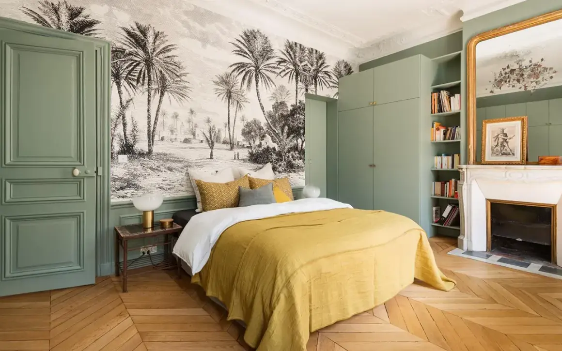 Une chambre adulte avec un papier peint paysage tropical, un lit jaune, une armoire verte et une cheminée.