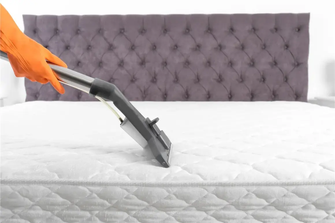 Main tenant en orange nettoie un matelas blanc avec un appareil à vapeur. Le nettoyage du matelas à la vapeur est essentiel pour une literie saine.