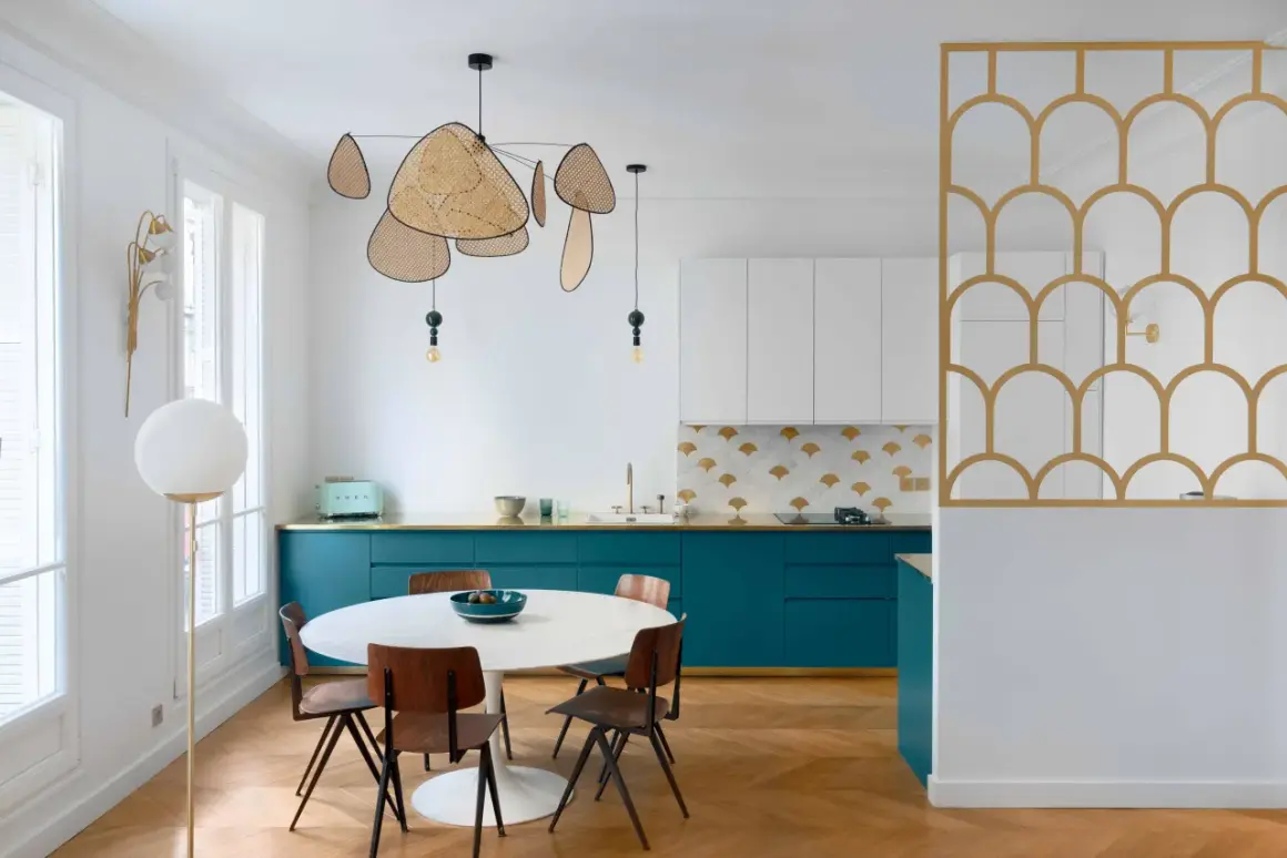 Cuisine moderne avec des armoires bleu canard, un dosseret à motifs dorés et blancs, et une table ronde. La composition de cuivre blanc se retrouve dans le luminaire aérien et la séparation décorative.