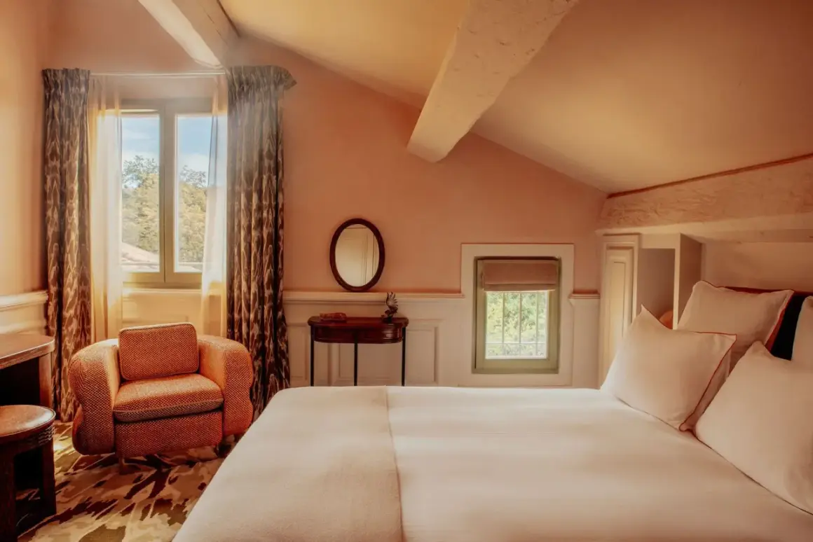 Chambre avec lit blanc, fauteuil orange, miroir rond et fenêtres donnant sur la verdure.