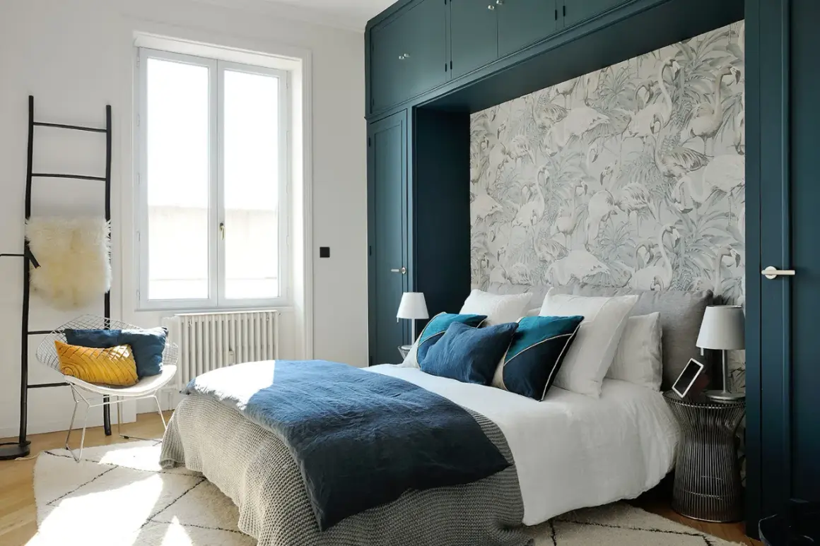 Chambre avec papier peint flamants roses, lit bleu marine et armoire vert foncé.