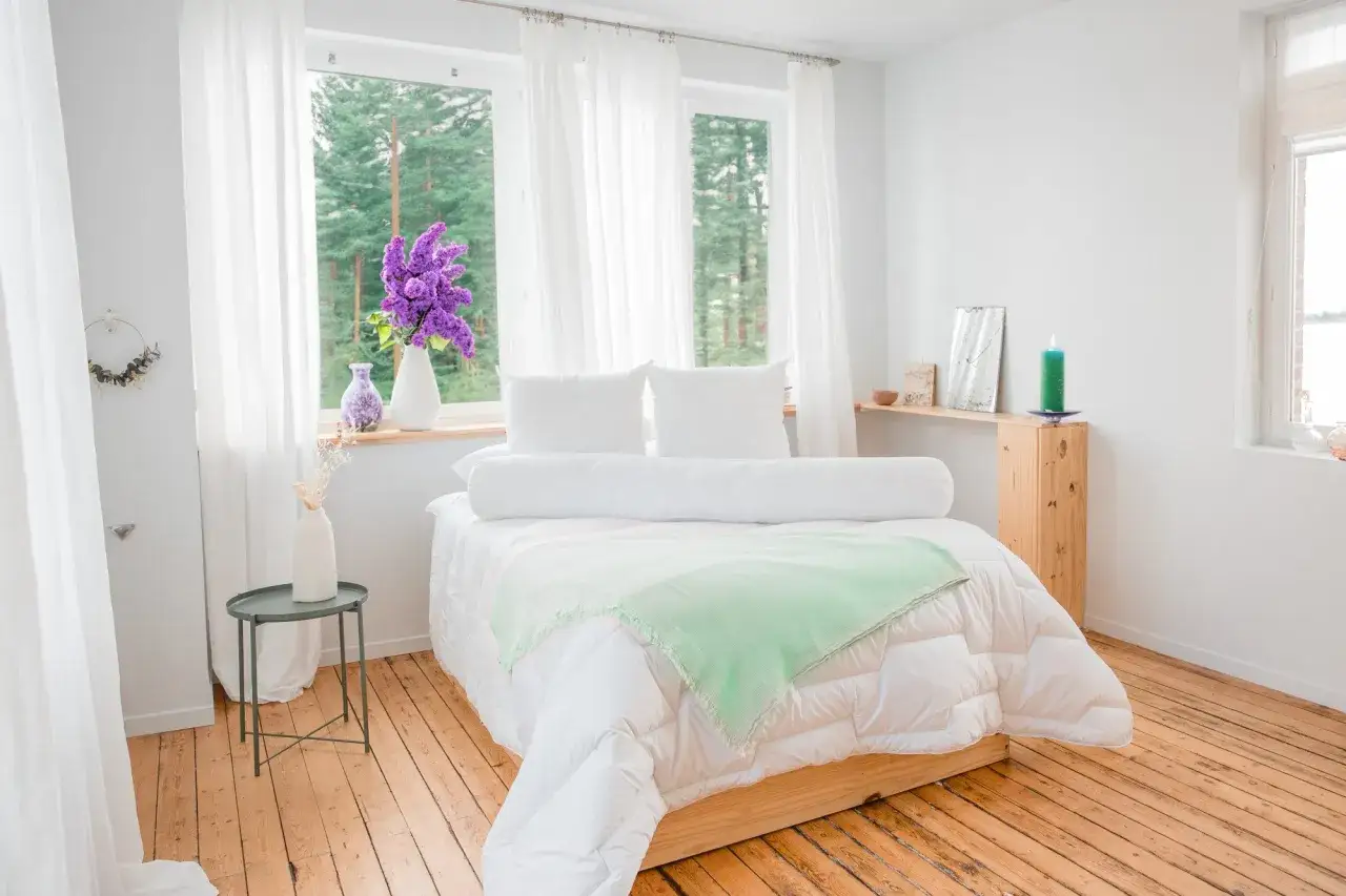 Un lit douillet avec un traversin blanc, des oreillers moelleux et une couverture verte. Des rideaux blancs et des fleurs violettes apportent une touche de fra&icirc;cheur.