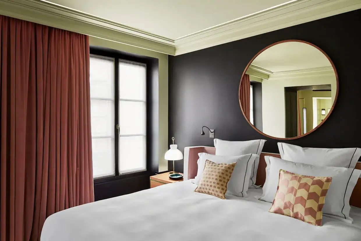 Un grand miroir rond au-dessus du lit reflète une chambre élégante avec des rideaux rouille et un mur sombre.