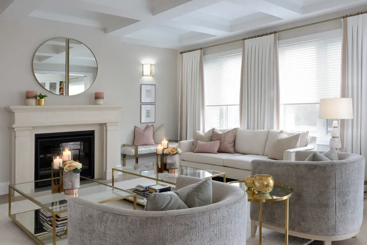 Salon lumineux avec cheminée en pierre, canapé crème, fauteuils gris et tables basses en verre. La couleur des métaux dorés rehausse les miroirs, les luminaires et les pieds des meubles.