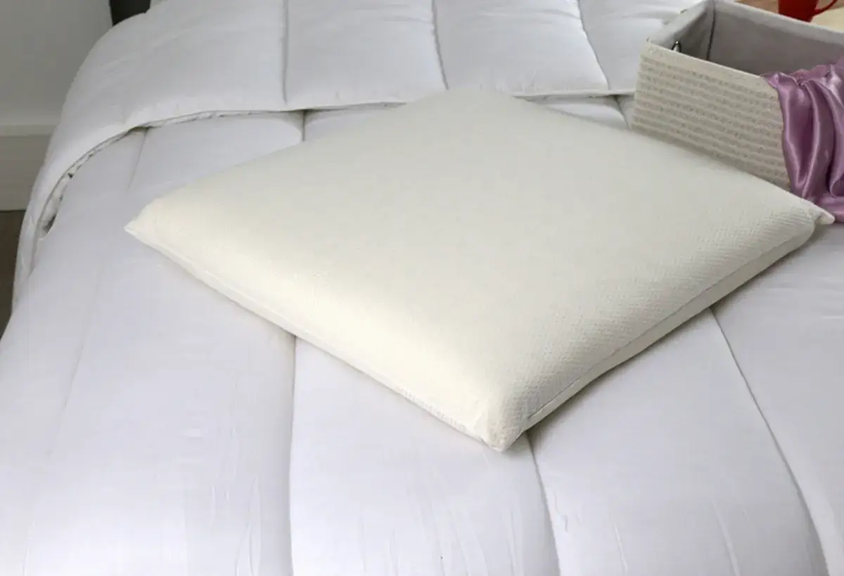 Un oreiller m&eacute;moire de forme blanc est pos&eacute; sur une couette blanche. Un panier en osier avec du tissu violet est &agrave; c&ocirc;t&eacute;.