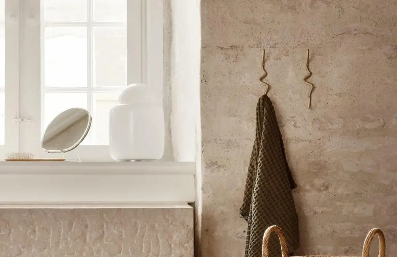 Deux patères en couleur laiton aux formes ondulées accueillent une serviette verte. Un miroir et un pot blanc sont posés sur une étagère.