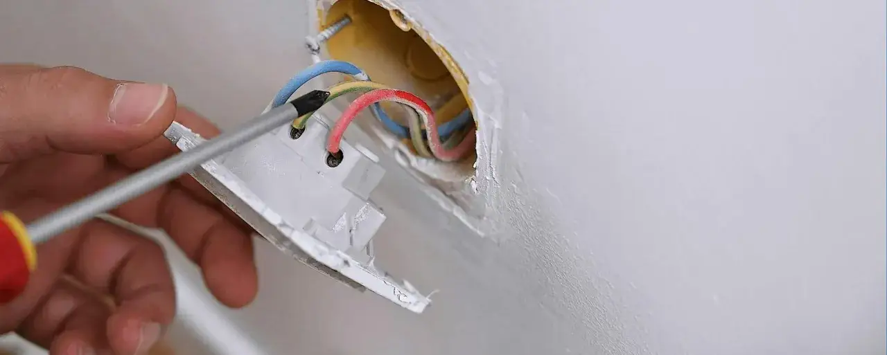 Main d'un &eacute;lectricien effectuant le branchement fiche &eacute;lectrique 3 fils. Les fils rouge, bleu et vert/jaune sont visibles.