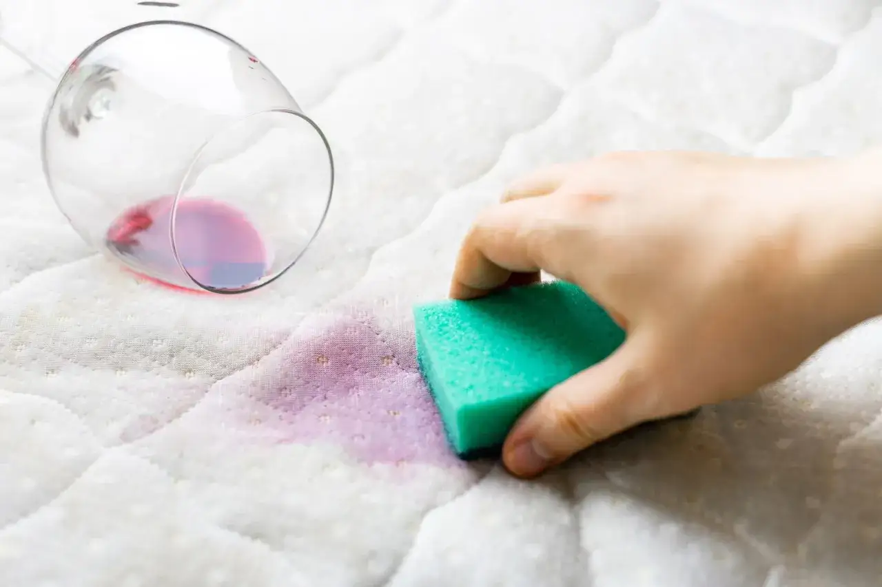 Main tenant nettoie un matelas jauni avec une &eacute;ponge verte, apr&egrave;s qu'un verre de vin rouge se soit renvers&eacute;.