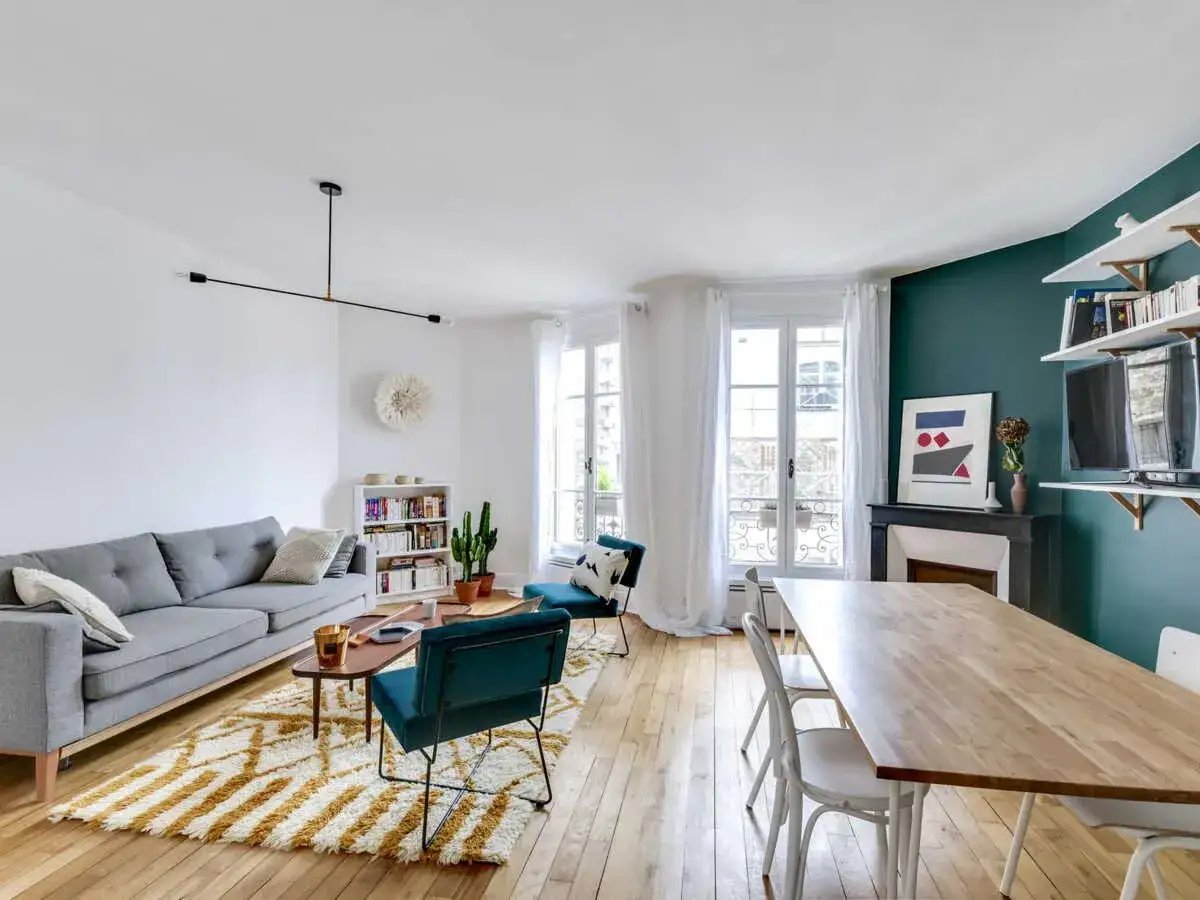 Un salon moderne et lumineux avec un canapé gris, un tapis graphique, des fauteuils en velours bleu canard et une table à manger en bois. La déco salon est complétée par une bibliothèque et des étagères murales.