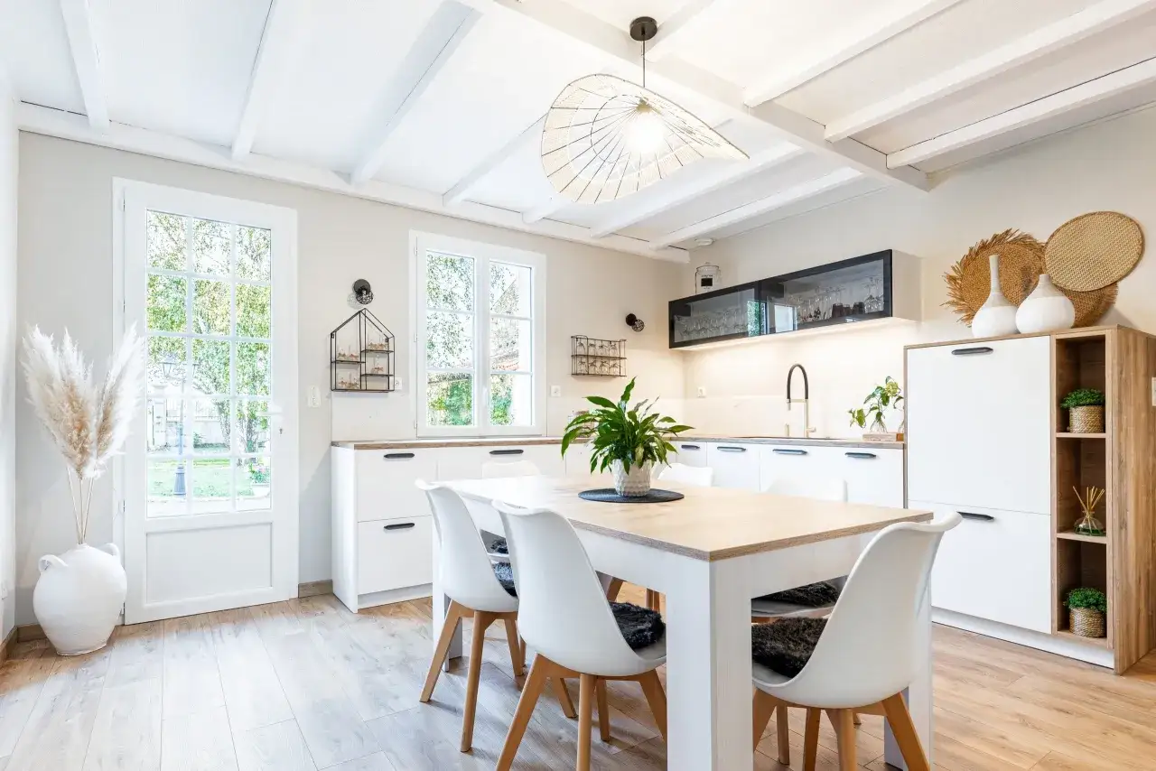 Une cuisine blanche style campagne chic, lumineuse avec poutres apparentes, table en bois, chaises modernes et une grande porte vitrée donnant sur le jardin.