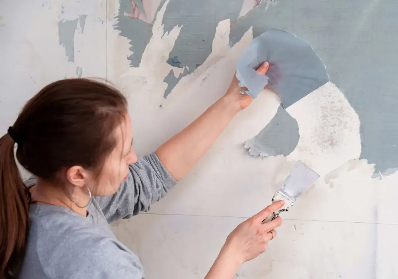 Une femme utilise une spatule pour d&eacute;coller du papier peint, une astuce de grand-m&egrave;re pour un d&eacute;collage facile.